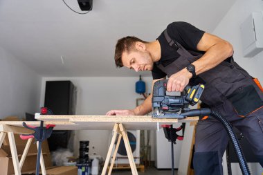 Marangoz ev atölyesinde elektrikli testereyle tahta keser. Garajda ahşap tezgahta çalışan bir adam. Ahşap işçiliği ve marangozluk kavramı