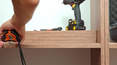 Mobilya montajı sırasında tahta dolabı mezurayla ölçen, arka planda aletleri ve vidaları olan bir adamın yakın çekimi. Ev yenileme ve DIY mobilya projesi kavramı