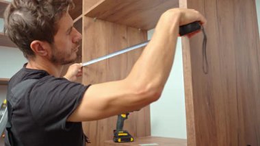 Mobilya montajı sırasında tahta dolabı mezurayla ölçen, arka planda aletleri ve vidaları olan bir adamın yakın çekimi. Ev yenileme ve DIY mobilya projesi kavramı