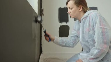 Koruyucu takım elbiseli bir kadın modern odada yenileme aşamasındaki fırçayla iç duvarı siyaha boyuyor.
