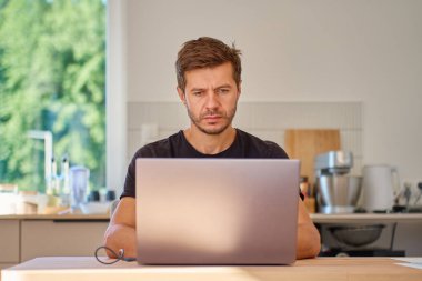 Parlak mutfaktaki ahşap masada dizüstü bilgisayarda çalışan bir adam. Ev ofisinde çalışan serbest çalışan biri. Uzak çalışma ve çevrimiçi iş kavramı