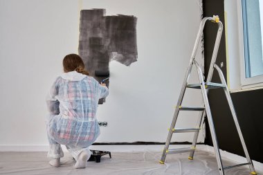 Koruyucu takım elbiseli, beyaz pantolonlu, siyah baskılı, yenileme sırasında tekerlekli paten kullanan bir kadın. İç duvarı boyayan kadın işçi. Evde tadilat ve iyileştirme kavramı