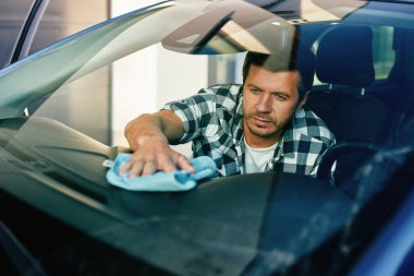 Adam arabanın içinde otururken arabanın ön panelini mikrofiber bezle siliyor. İç temizlik, araç bakımı ve araba bakımı kavramı