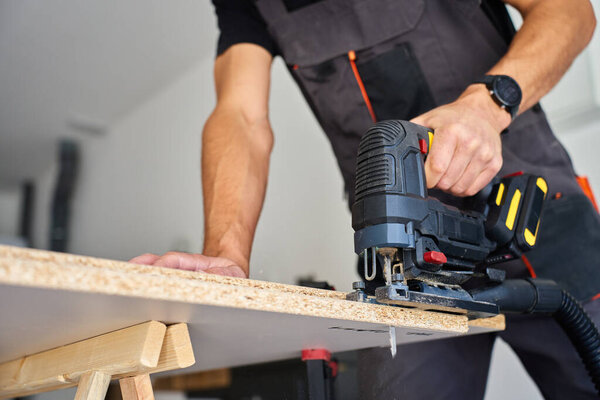 Carpenter cuts wooden board with electric jigsaw in home workshop. Man in workwear working with wood on workbench in garage. Woodworking and carpentry concept