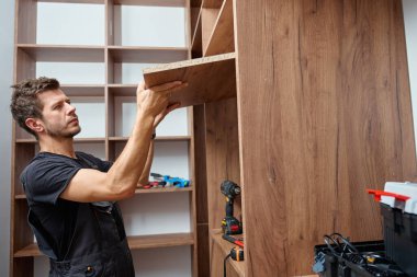 Evdeki ahşap gardırobu, aletleri kullanarak ve büyük ahşap paneli tutarak yapan adam. DIY mobilya montajı, ev geliştirme ve yenileme işi kavramı