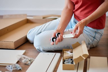 Yerde oturan bir kadın, elinde tekerlekler ve aletlerle yatak parçaları birleştiriyor. DIY mobilya montajı, ev geliştirme ve kendi kendini kurma süreci kavramı