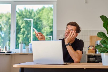 Adam evde dizüstü bilgisayarla çalışıyor ve klimanın ısısını ayarlamak için uzaktan kumanda kullanıyor. Ofiste çalışırken sıcakla boğuşan kişi