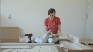 Mobilya montajı sırasında yerde oturmuş kullanma kılavuzunu okuyan bir kadın. Yerde aletlerin ve yeni yatağın parçalarının yanında oturan biri. DIY ev geliştirme ve mobilya kurulumu kavramı