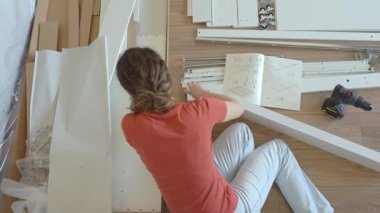 Kadın ahşap zeminde oturuyor ve yeni evdeki mobilyaları monte ediyor, beyaz yatak çerçevesinin parçalarını bağlıyor. DIY ev yenileme ve taşınma konseptinden sonra ev organizasyonu