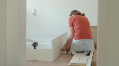 Kadın ahşap zeminde oturuyor ve yeni evdeki mobilyaları monte ediyor, beyaz yatak çerçevesinin parçalarını bağlıyor. DIY ev yenileme ve taşınma konseptinden sonra ev organizasyonu