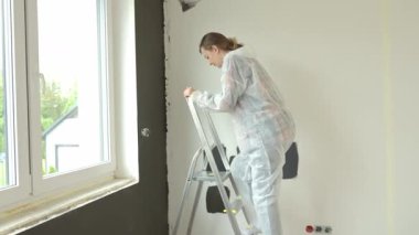 Koruyucu giysili kadın elinde boya silindiriyle merdiveni tırmanıyor ve siyah aksan duvarını boyamaya hazırlanıyor. Modern apartman içinde ev yenileme