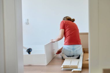 Kadın ahşap zeminde oturuyor ve yeni evdeki mobilyaları monte ediyor, beyaz yatak çerçevesinin parçalarını bağlıyor. DIY ev yenileme ve taşınma konseptinden sonra ev organizasyonu