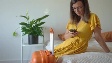 Sarı elbiseli kadın turuncu bavullu yatakta oturuyor ve akıllı telefon kullanıyor. Dişi gezgin, yeşil bitkilerle modern iç mekanda ayrılmayı bekliyor. Seyahat ve yolculuk hazırlığı kavramı