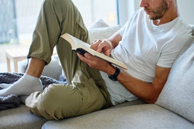 Kanepede uzanmış, günlük giysiler içinde kitap okuyan bir adamın yakın çekimi. Rahatlama, rahatlama ve kendini eğitme konsepti