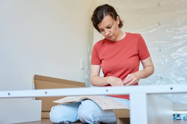 Kadın ahşap zeminde oturuyor ve yeni evdeki mobilyaları monte ediyor, beyaz yatak çerçevesinin parçalarını bağlıyor. DIY ev yenileme ve taşınma konseptinden sonra ev organizasyonu