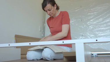 Kadın ahşap zeminde oturuyor ve yeni evdeki mobilyaları monte ediyor, beyaz yatak çerçevesinin parçalarını bağlıyor. DIY ev yenileme ve taşınma konseptinden sonra ev organizasyonu