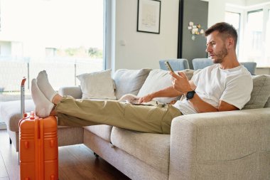 Beyaz gömlekli adam turuncu bavulun yanında gri koltukta oturuyor ve akıllı telefon kullanıyor. Erkek gezgin modern oturma odasında köpekle evden ayrılmayı ya da seyahat etmeyi bekliyor.