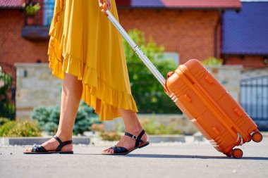 Sarı elbiseli, turuncu valizli bir kadın güneşli bir günde sokakta yürüyor. Bagajlı kadın gezgin yolculuktan evine dönüyor.