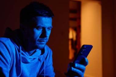 Kapüşonlu bir adam geceleri cep telefonu kullanıyor. Yüzünde mavi ışık olan karanlık bir odada akıllı telefon ekranına bakıyor. Teknoloji kullanımı, telefon bağımlılığı kavramı