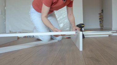 Kadın ahşap zeminde oturuyor ve yeni evdeki mobilyaları monte ediyor, beyaz yatak çerçevesinin parçalarını bağlıyor. DIY ev yenileme ve taşınma konseptinden sonra ev organizasyonu