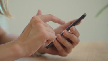 Masada oturan ve modern akıllı telefon kullanan bir kadının yakın çekimi. Kadın eli cep telefonu tutuyor, sosyal medyada geziniyor veya evde mesajlaşıyor