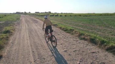 Cyclist wearing helmet and sportswear riding gravel bike on a dirt road near green farmland. Man training on bicycle. Active lifestyle concept