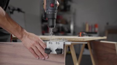 Close-up of carpenter drilling hole in wooden panel using drill and metal jig tool for precision. Man working in home workshop. Concept of woodworking, furniture making and DIY home projects