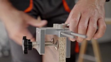 Close-up of carpenter drilling hole in wooden panel using drill and metal jig tool for precision. Man working in home workshop. Concept of woodworking, furniture making and DIY home projects