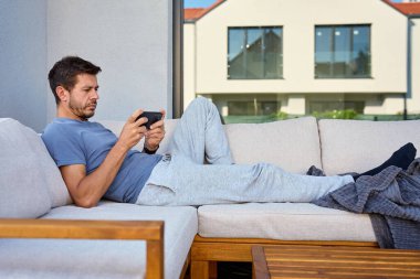 Yetişkin bir adam, günlük giysiler içinde kanepede uzanıyor ve açık havada akıllı telefondan oyun oynuyor. Terasta rahatlayan, cep telefonu kullanan biri. Dijital yaşam tarzı, eğlence ve çevrimiçi eğlence anlayışı