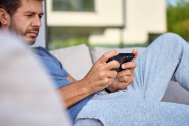 Dışarıdaki ahşap sandalyede oturan, akıllı telefonu elinde tutan sıradan elbiseli bir adam. Terasta cep telefonuyla oyun oynayan kişi. Rahatlama, çevrimiçi oyun ve dijital yaşam tarzı kavramı