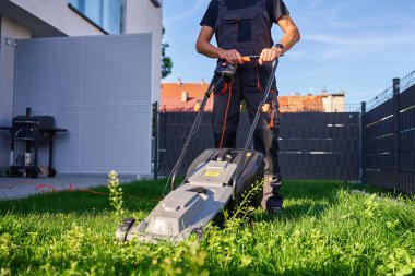 Arka bahçede çim biçmek için elektrikli çim biçme makinesi kullanan biri. Banliyö evinin yakınında çim biçen bir adam. Bahçe bakımı, bahçe bakımı ve açık hava işi kavramı