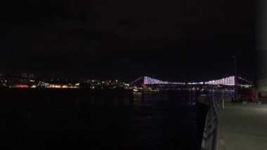 Hava karardıktan sonra İstanbul 'da Uskudar kıyısı boyunca yürümek. 15 Temmuz Şehitler Köprüsü görünür ve karanlık sulara yansıyan parlak bir ışık..