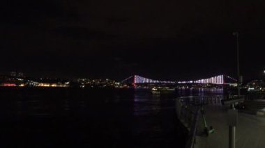 Hava karardıktan sonra İstanbul 'da Uskudar kıyısı boyunca yürümek. 15 Temmuz Şehitler Köprüsü görünür ve karanlık sulara yansıyan parlak bir ışık..