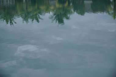 Su sakin ve gökyüzü bulutlu. Sudaki ağaçların yansıması barışçıl bir atmosfer yaratır.