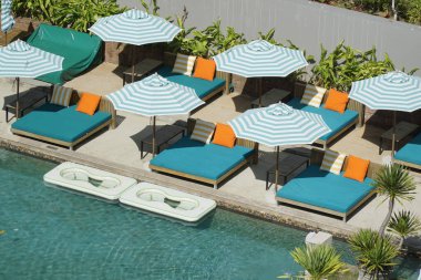 Çeşitli mavi ve beyaz şemsiyeli ve şezlonglu havuz. Havuz beyaz bir çitle çevrili ve yakınlarda birkaç saksı bitkisi var.