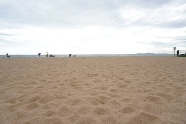 Kumsalda birkaç kişi oturuyor. Gökyüzü bulutlu ve kumsal boş.