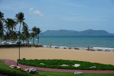 Plaj sahnesi, palmiye ağaçları ve güneşin tadını çıkaran insanlar. Sahil insanlarla dolu ve gölge için kurulmuş birçok şemsiye var. Okyanus sakin ve gökyüzü berrak. Huzurlu bir hava yaratıyor.