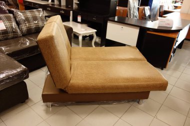 Kahverengi aksanlı bronz kanepe bir mobilya mağazasında sergileniyor. Kanepe bir fayans zeminine yerleştirilir ve sandalyeler ve masalar da dahil olmak üzere diğer mobilyalarla çevrilidir. Mağazanın modern bir