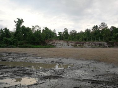 Arka planda tepe olan çamurlu bir alan. Gökyüzü bulutlu ve güneş batıyor.