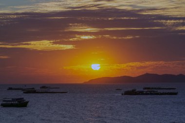 Güneş okyanusun üzerinde batıyor, suyun üzerine sıcak bir parıltı saçıyor. Gökyüzü bulutlarla dolu, sakin ve huzurlu bir atmosfer yaratıyor. Sudaki tekneler dağıtıldı.