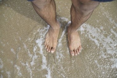 İnsanın ayakları suyun içinde, ve su köpüklü. Adam sahilde duruyor ve su sakin.