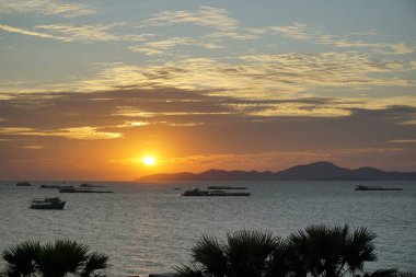 Güneş okyanusun üzerinde batıyor, suyun üzerine sıcak bir parıltı saçıyor. Gökyüzü bulutlarla dolu, sakin ve huzurlu bir atmosfer yaratıyor. Sudaki tekneler etrafa saçıldı.