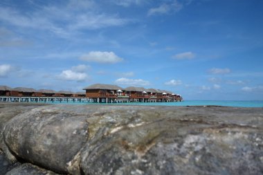 Küçük ahşap evlerle dolu bir sahil. Evlerin hepsi aynı renkte ve sahil boyunca dizilmiş. Gökyüzü mavi ve arka planda bazı bulutlar var