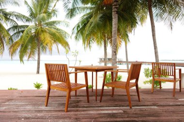 Tahta masa ve sandalyeler plaja kurulmuş. Sandalyeler masanın etrafına dizildi ve arka planda palmiye ağaçları var. Sahne huzurlu ve rahatlatıcı.