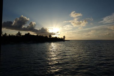 Güneş okyanusun üzerinde batıyor, suyun üzerine sıcak bir parıltı saçıyor. Gökyüzü bulutlarla dolu. Huzurlu ve huzurlu bir atmosfer yaratıyor.