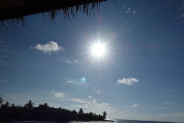 Güneş gökyüzünde ışıl ışıl parlıyor, okyanusun üzerine sıcak bir parıltı saçıyor. Bulutlar gökyüzüne dağılmış, derinlik ve hareket hissi katıyor.