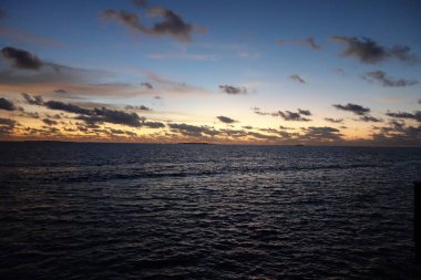 Gökyüzü karanlık ve bulutlu ve okyanus sakin. Güneş batıyor ve su gökyüzünün renklerini yansıtıyor.