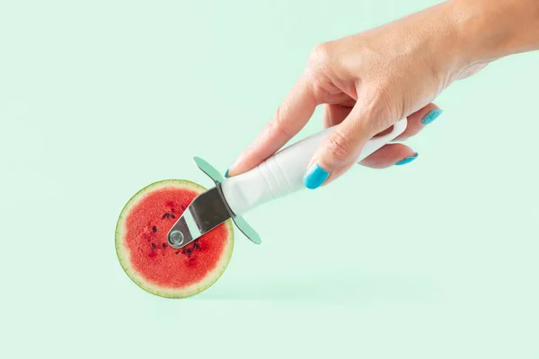 Womans hand holding pizza cutter with watermelon slice on isolated pastel green background. Minimal abstract concept of summer fruit, healthy food, eating habits, boost immunity and hydration.