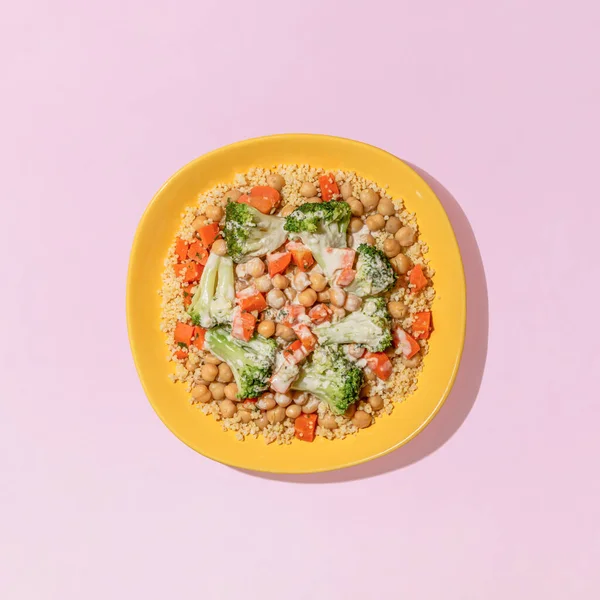 Yellow plate with vegan Buddha bowl with millet, broccoli, carrots, crunchy chickpeas, all topped with delicious garlic tahini dressing. Pink background with shadow. Minimal flat lay. Food concept.