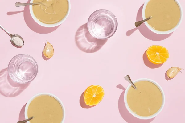 Halved lemon, tahini, garlic, salt and glasses of water on isolated pastel pink background with shadows and reflections. Aesthetic food concept. Flat lay. Minimal creative tahini sauce pattern.